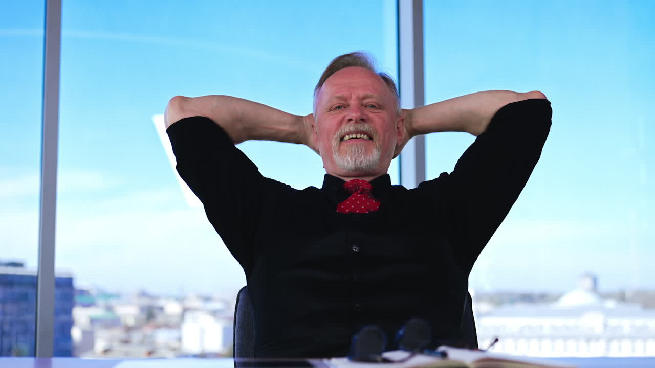 Senior Caucasian man sits at desk holding his head in hands. Man leans back in the chair putting his hands behind the head and smiles. Panoramic window at backdrop.