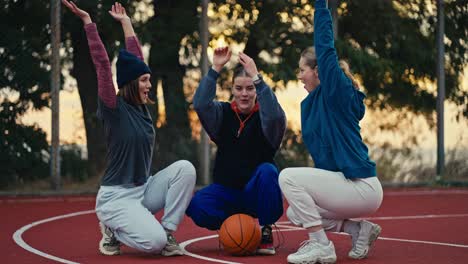 Three-happy-blonde-girls-in-sportswear-put-their-hands-together-and-raise-them-up-during-the-start-of-a-basketball-match-on-a-red-sports-court-in-the-morning