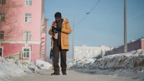 cinematographer wearing winter coat and black shoes adjusts camera lens while walking along snow covered walkway under bright sun in busy urban city with passing cars and tall buildings