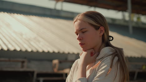 thoughtful young woman outdoors, wearing hoodie, gently holding her chin as she walks in a quiet stadium with blurred background of a roof, bathed in soft sunlight