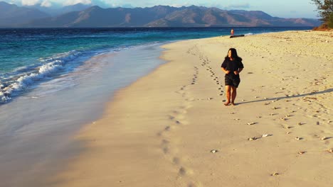 Beautiful-young-woman-taking-a-casual-stroll-in-the-soft-sand-of-a-tropical-island-with-the-afternoon-sun-setting