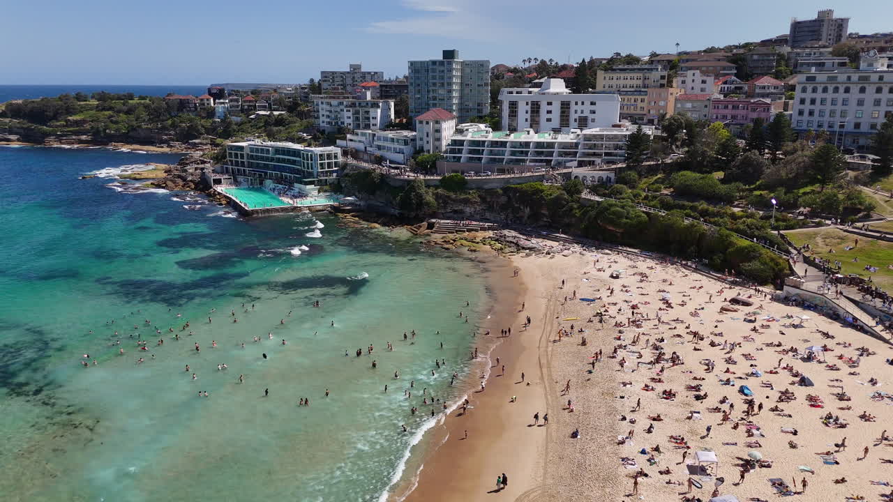 Aerial footage of Bondi Beach, featuring beach lovers enjoying the golden sand and turquoise waters. New South Wales, Australia. Wide drone footage.