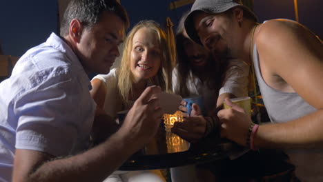 group of friends sitting in the evening on the street for a table one of the men shows photos on his smartphone the woman laughs