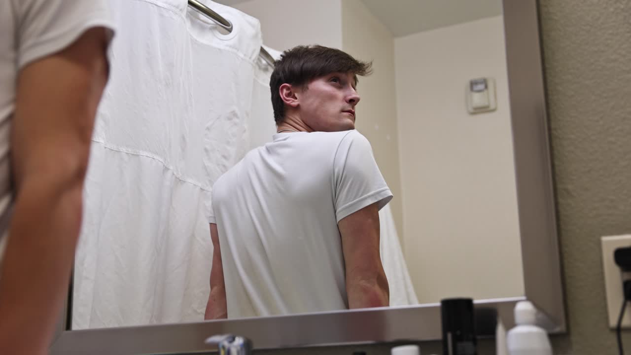 젊은 백인 남성이 호텔 방의 욕실 거울을 향해 자신 있게  ⁇ 고 과거를 회상하고 있습니다.