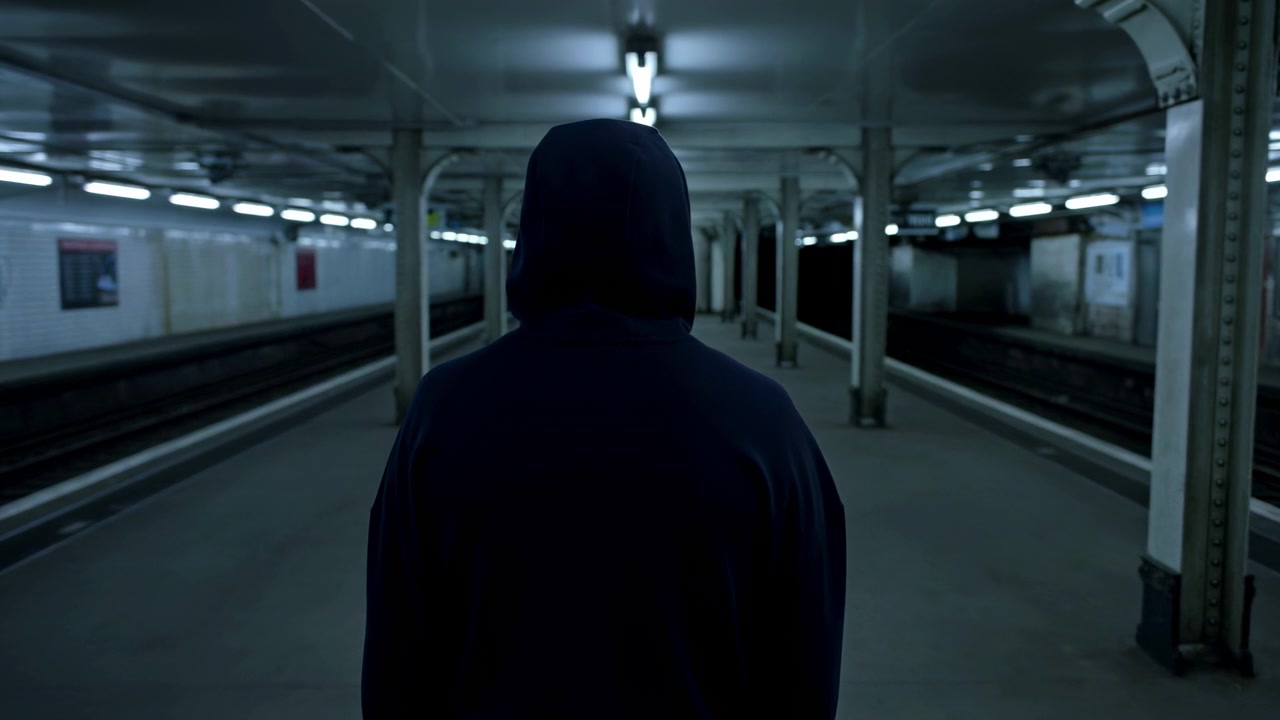 Vista posteriore di una figura incappucciata rimane centrata mentre la prospettiva si restringe su una tranquilla piattaforma della stazione, con illuminazione dall'alto fredda, binari paralleli, pareti piastrellate e colonne ripetute.