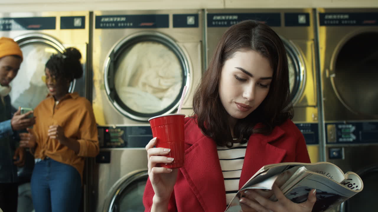 jovencita bonita sentada en el cuarto de servicio de lavandería y leyendo una revista de moda mientras bebe con pajita