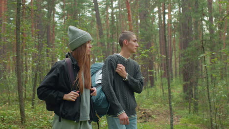close up of siblings walking side by side through forest, girl adjusting backpack straps while boy grips sleeping bag strap over shoulder, both dressed casually and surrounded by trees