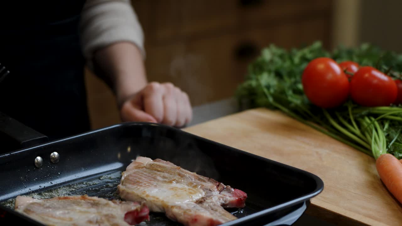Pork chops cook on grill pan near carrots and tomatoes in rustic kitchen
