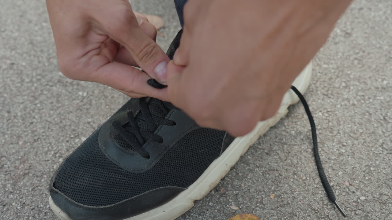 Corridore che si prepara le scarpe, individuo atletico che si allaccia le scarpe all'aperto, persona che si allaccia scarpe nere all'aperto al mattino, primo piano delle mani che allacciano le scarpe da ginnastica su una superficie pavimentata durante la corsa mattutina