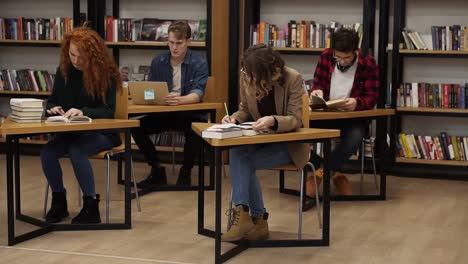 group of students preparing for exam in university library, two beautiful females making notes, two mates behind reading book, working on laptop, concentrated students in studying process