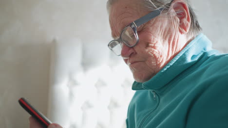elderly woman wearing turquoise fleece sweater, adjusting glasses while holding smartphone in one hand. focused expression, sunlight softly illuminating her face and surroundings