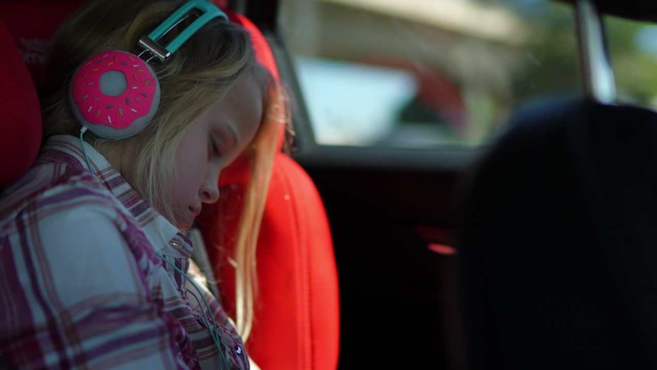 Young blonde girl enjoying music through donut shaped wireless headphones while sitting comfortably in passenger seat during family road trip