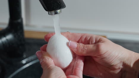 close-up of hands washing a white egg under a steady stream of running water from a black kitchen faucet, the person is wearing a plaid garment, emphasizing cleanliness, hygiene