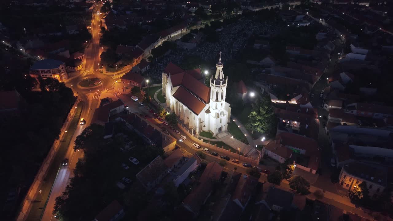Aerial view of a historic church brightly lit against the dark surroundings, with winding city streets illuminated by streetlights. The warm glow of the lights contrasts with the deep blue night sky