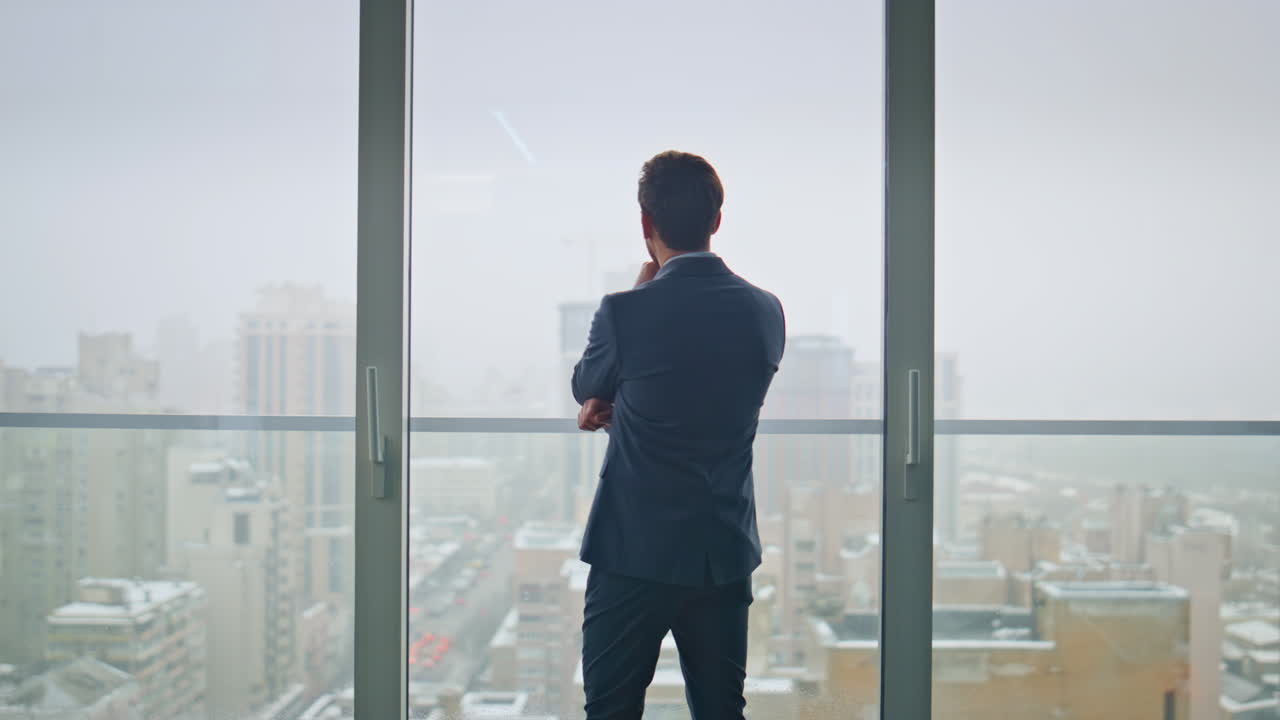 Uomo d'affari pensieroso di spalle che osserva lo skyline dalla finestra panoramica dell'ufficio.