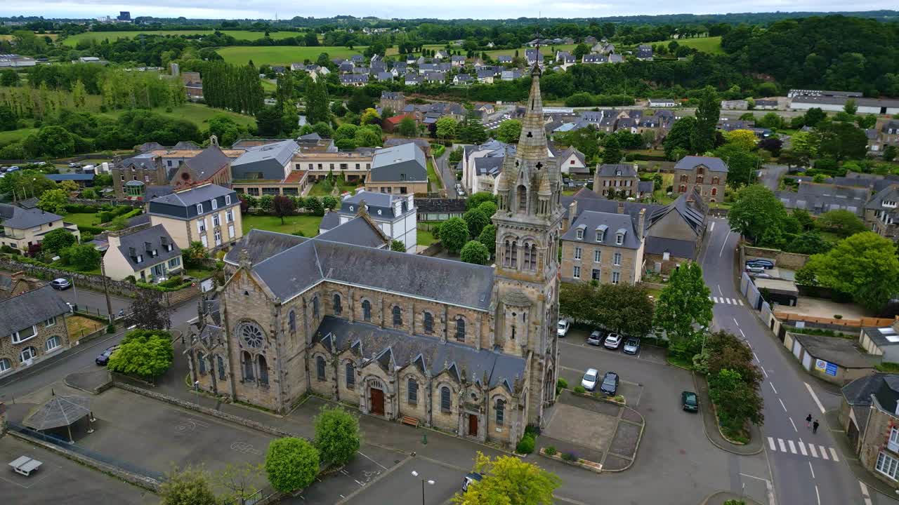 The drone flies laterally past the rose window side of Saint-Sauveur Church in Plancoët, with parked cars, nearby houses and green surroundings. Brittany in France