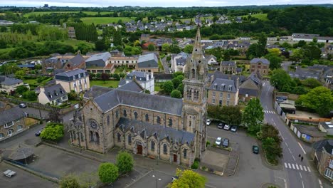 the drone flies laterally past the rose window side of saint-sauveur church in plancoët, with parked cars, nearby houses and green surroundings. brittany in france