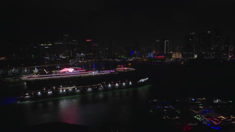Aerial-night-view-of-cruise-ship-in-city-downtown-port-against-the-background-of-illuminated-skyscrapers-skyline