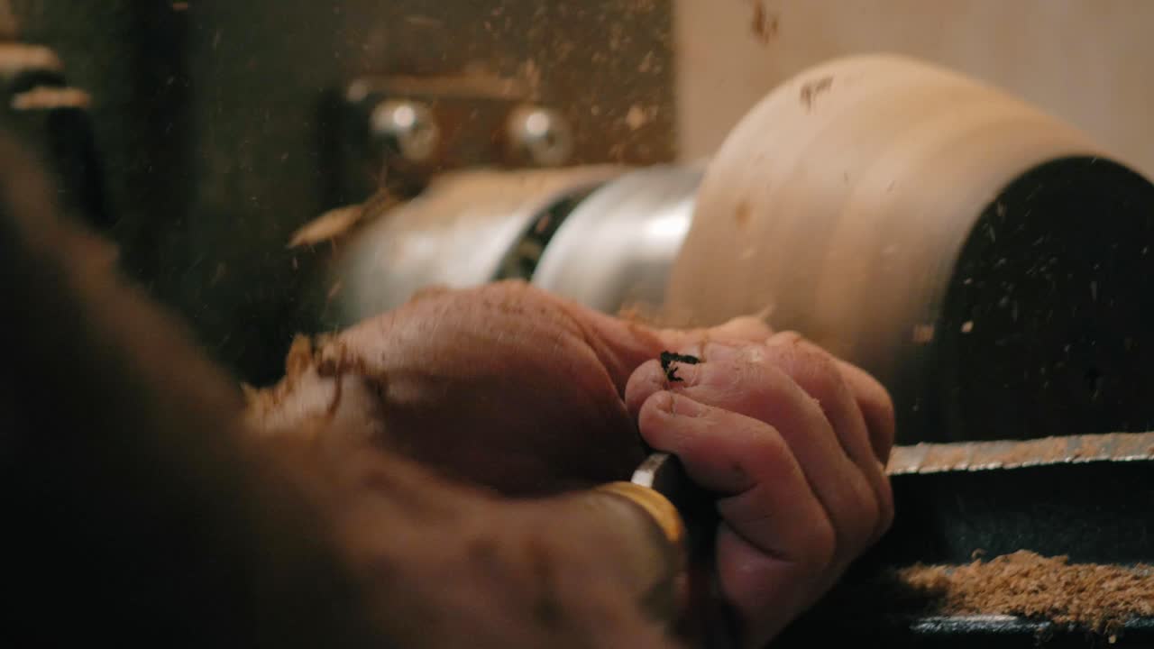 Artisan hands shape spinning wood on a lathe in a workshop. Flying shavings and sawdust surround the chisel during the carving process of traditional carpentry