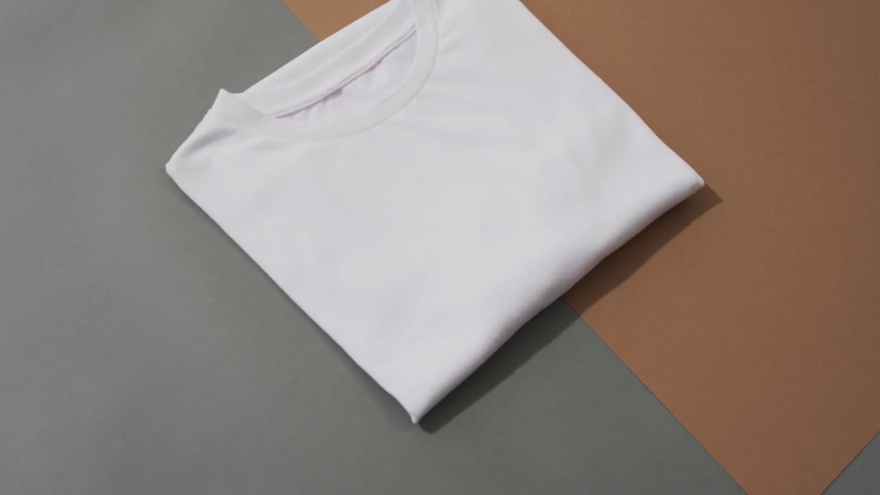 Video of flat lay of folded white t shirt with copy space on grey and brown background