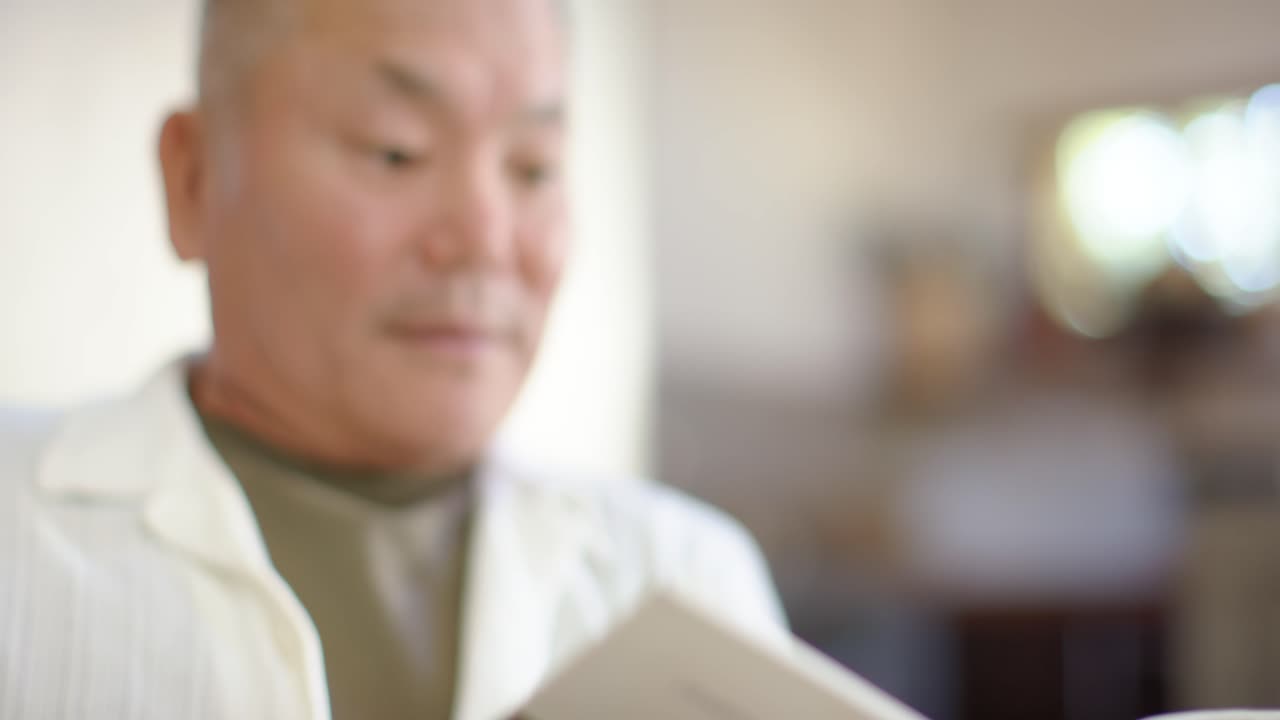 거실에서 페이퍼백 책 표지를 펼치고 손으로 페이지를 넘기며 글을 읽는 아시아계 노인 남성