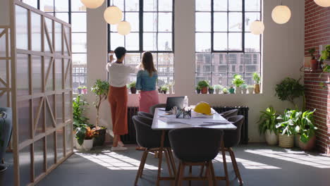 Mujeres-De-Negocios-Arquitectas-Discutiendo-El-Proyecto-De-Construcción-Equipo-De-Ingenieros-Intercambiando-Ideas-Planificando-Un-Desarrollo-Exitoso-Disfrutando-De-La-Vista-De-La-Ventana-En-Una-Oficina-Moderna