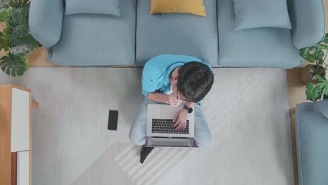 top view zoom out shot of a man thinking about something then raising index finger while typing on a laptop on the floor with smartphone in front of a sofa at home