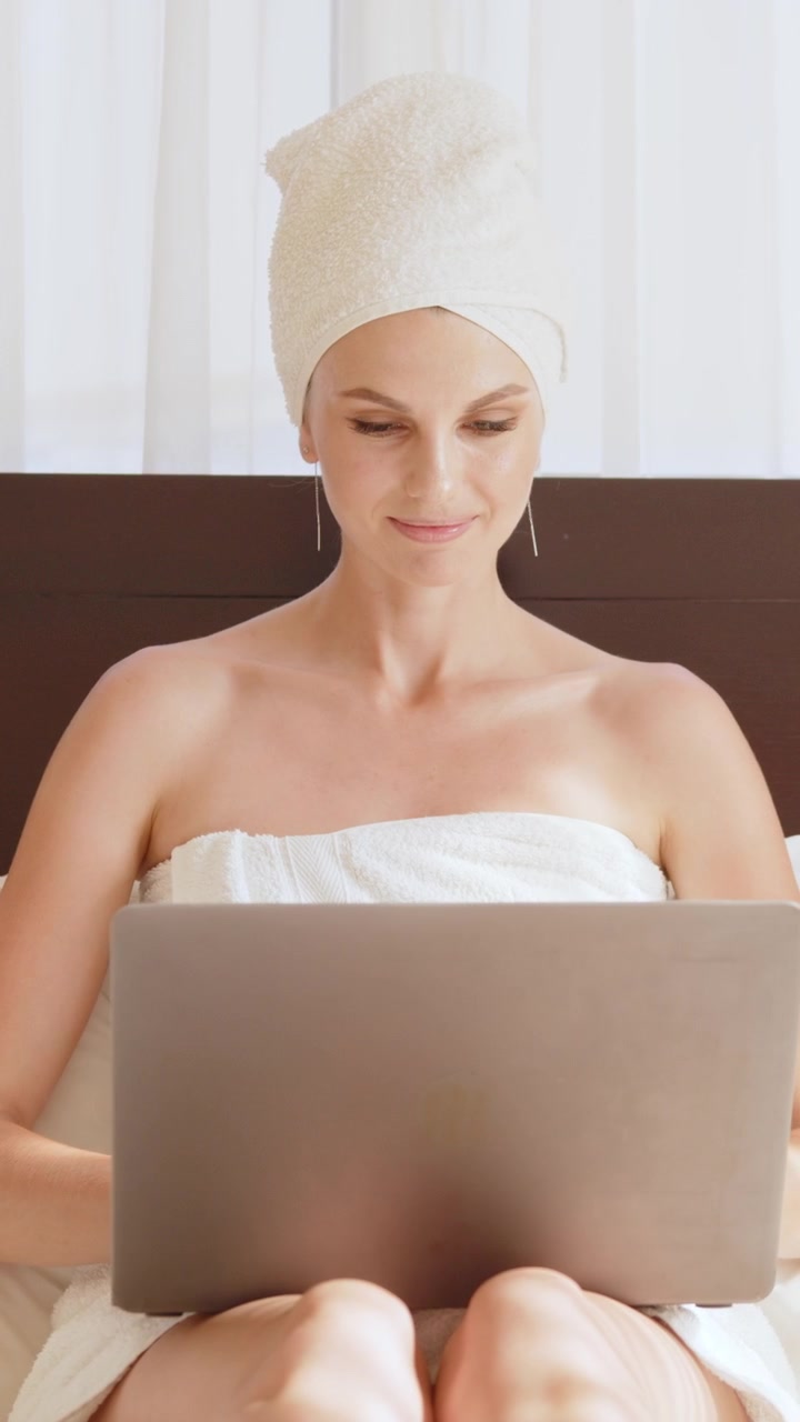 Una donna in asciugamano usa un laptop mentre si rilassa a letto, video verticale