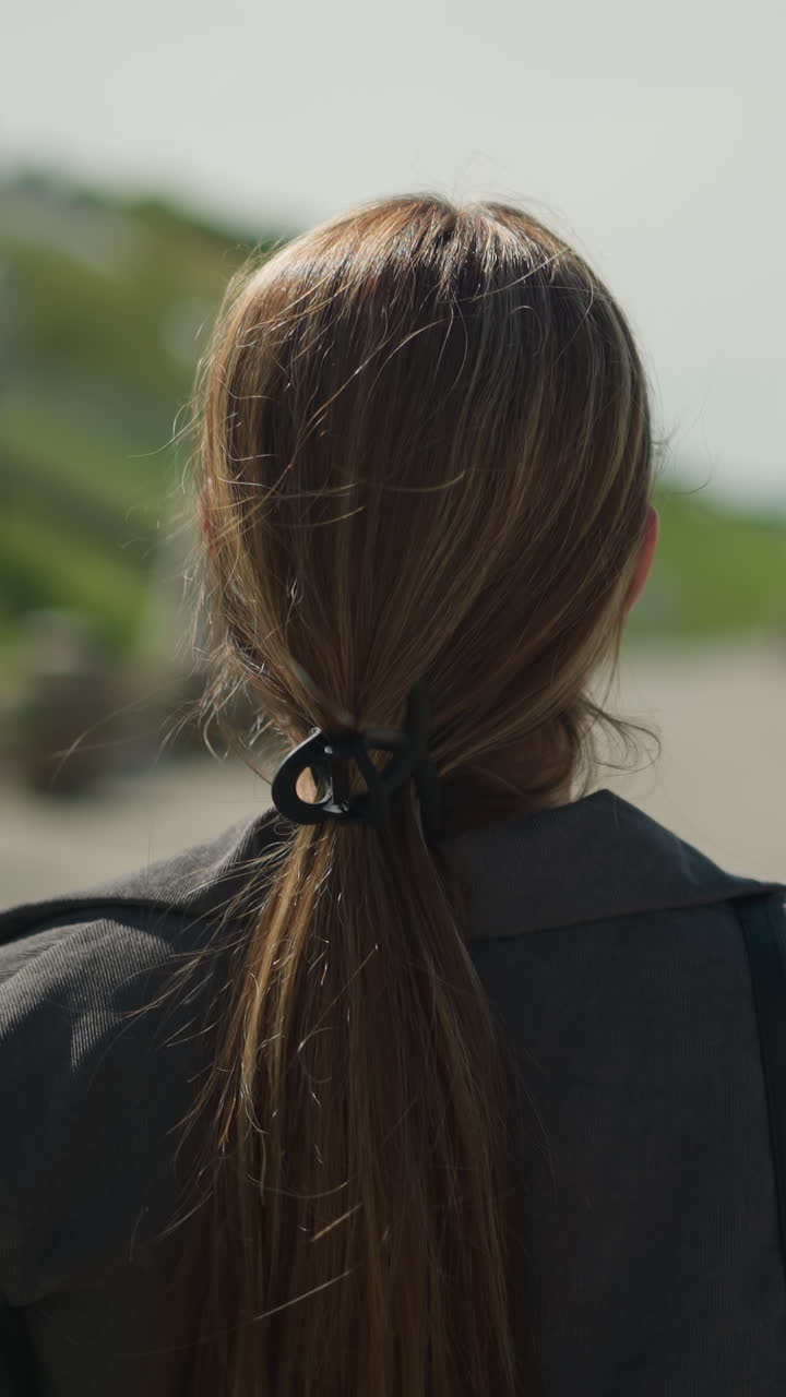 머리카락이 뒤로 여있는 타르로 된 도로를 고있는 여자의 뒷면, 멀리 왼쪽으로 바라보고, 배경은 약간 흐릿하고, 초록색과 도로 요소가 보입니다.