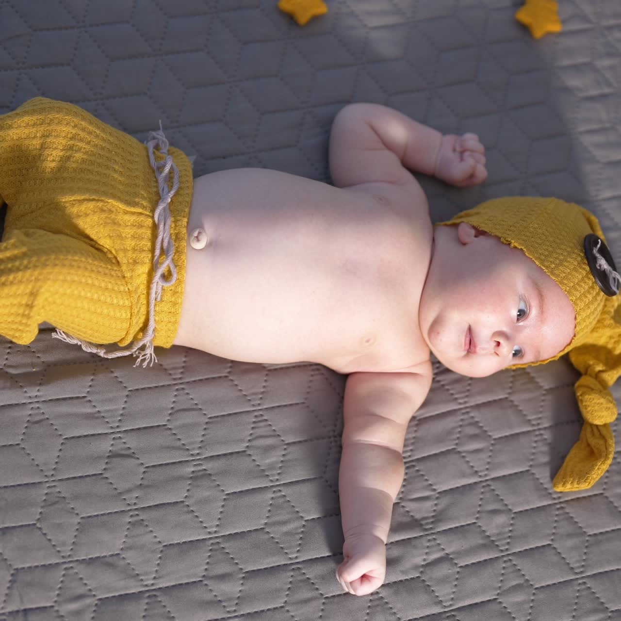 Kid in yellow hat with big button and wide pants is on the bed. Toddler in a funny suit with naked belly resting on a big bed. View from above