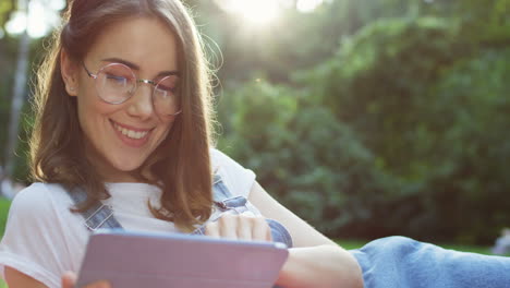 close-up view of a young caucasian woman in glasses watching something on the tablet while lying and resting on the grass in the park