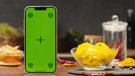 a modern greenscreen mobile device is displayed on a wooden table next to a bowl of food, demonstrating its versatility for multimedia projects