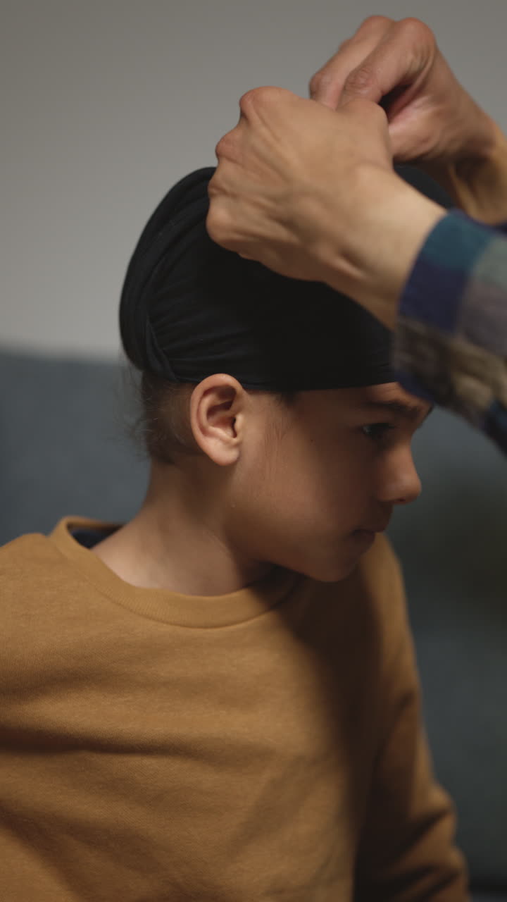 video vertical de primer plano de un padre atando un turbante a la cabeza de un joven sikh, su hijo con el nudo superior sentado en el sofá en casa 1
