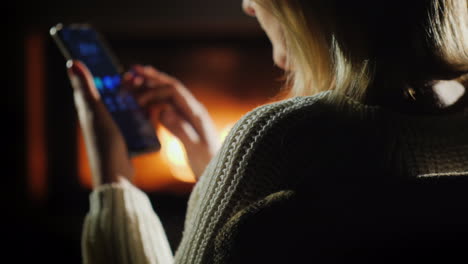 una mujer usa un teléfono inteligente por la noche sentada junto a la chimenea