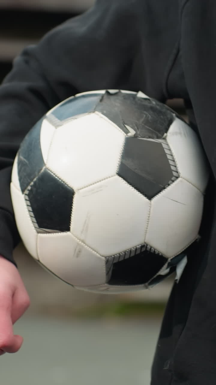 Close-up of someone holding a well-used soccer ball under the arm, with a blurred staircase in the background