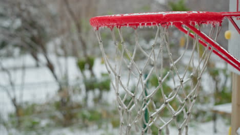 primer plano de un borde de aro de baloncesto rojo cubierto de hielo, red brillante con hielo, contra un telón de fondo de jardín nevado, capturando el agarre del invierno en equipos deportivos al aire libre