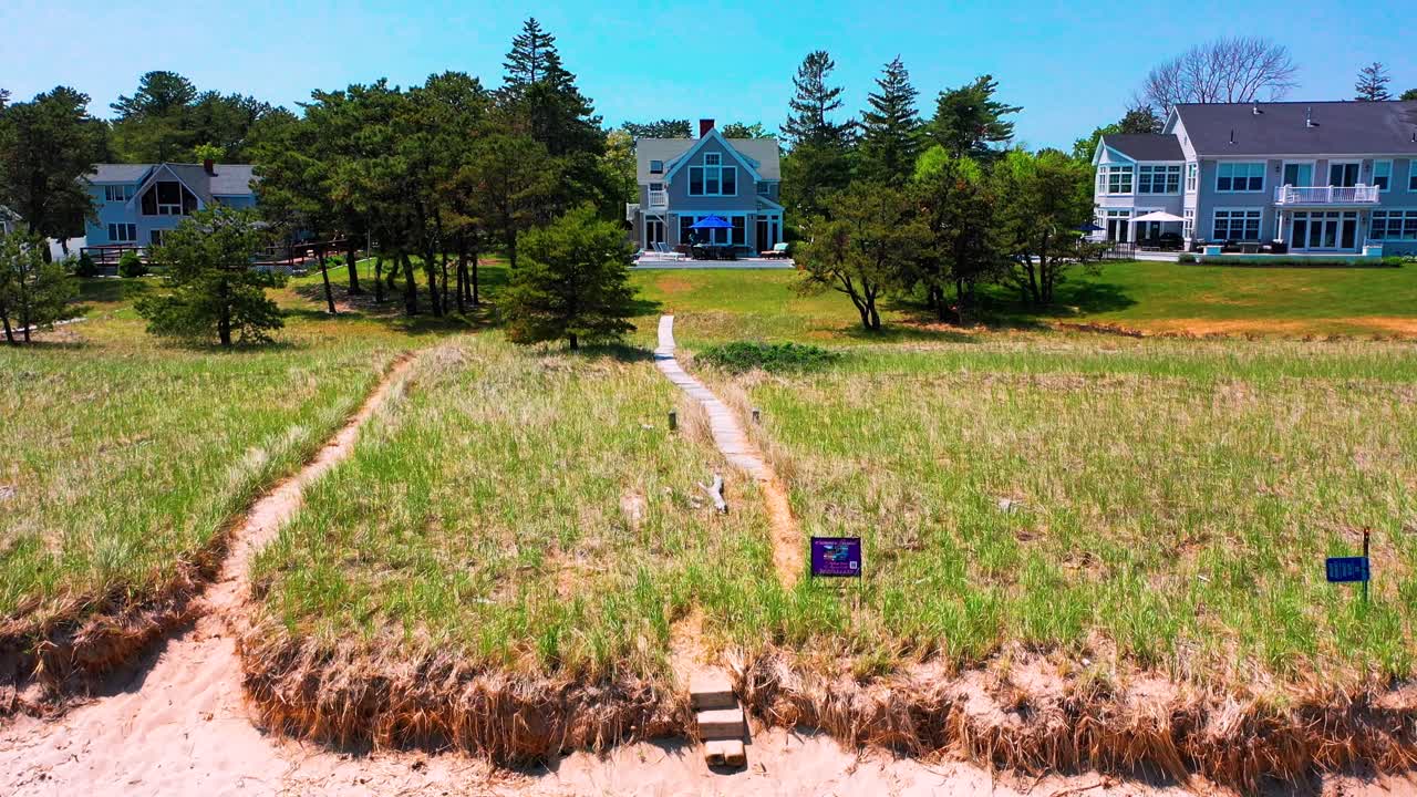 Point of view aerial drone flying over a wooden walkway bridge winding through tall grasses and trees, leading to the sandy Atlantic beach in Saco, Maine. A tranquil summer coastal outdoor vacation.