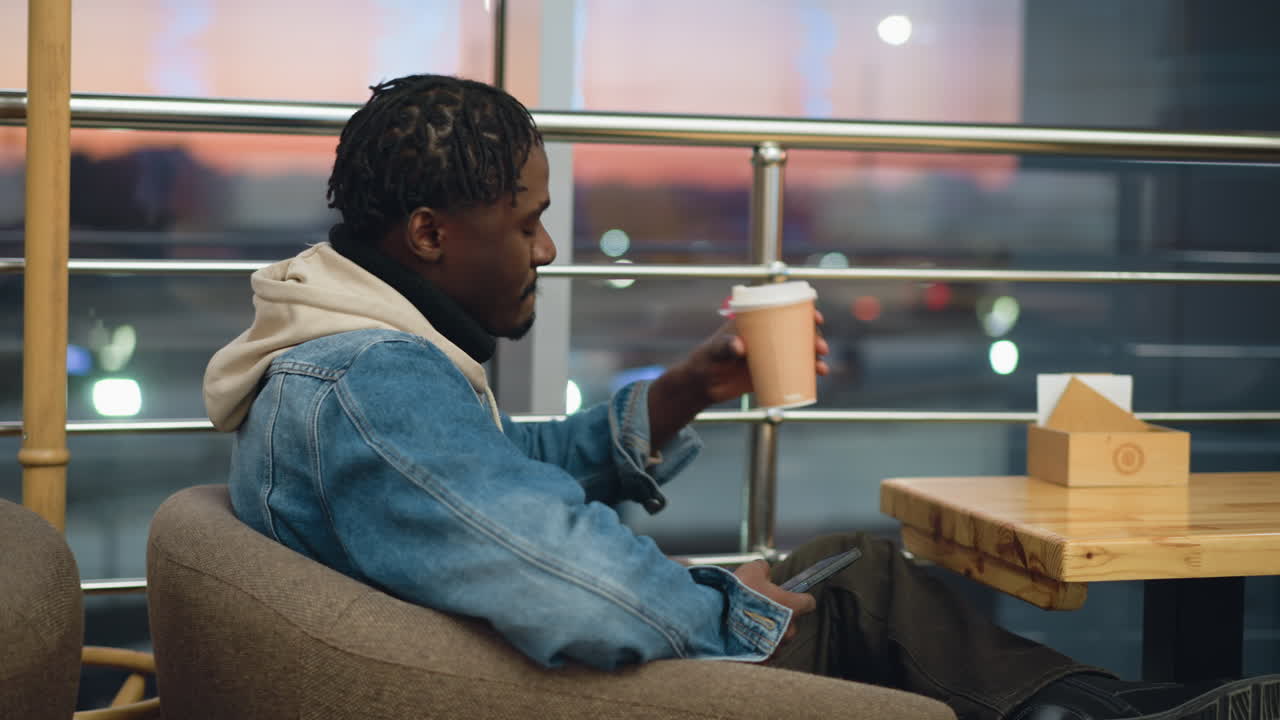 Guy in denim jacket and beige hoodie sitting in armchair at wooden table holding coffee cup, quietly enjoying serene indoor setting with glowing sunset and blurred city lights outside window