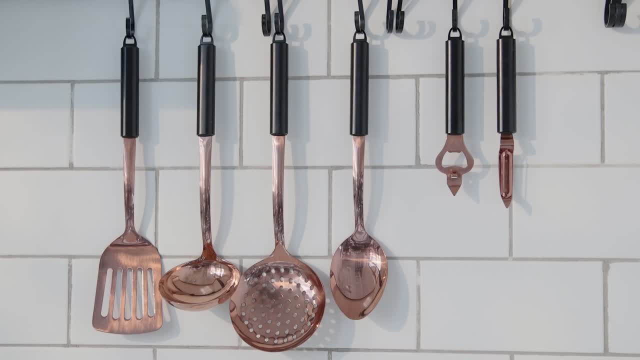 Copper kitchen utensils hang neatly on a tiled white wall, catching natural light in a minimal, modern cooking space.