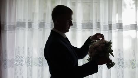 Handsome-groom-fixes-his-wedding-bouquet-near-window.-Silhouette