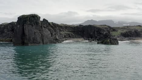 Low-flying-aerial-towards-small-beach-between-black-igneous-cliffs-of-Skardsvik