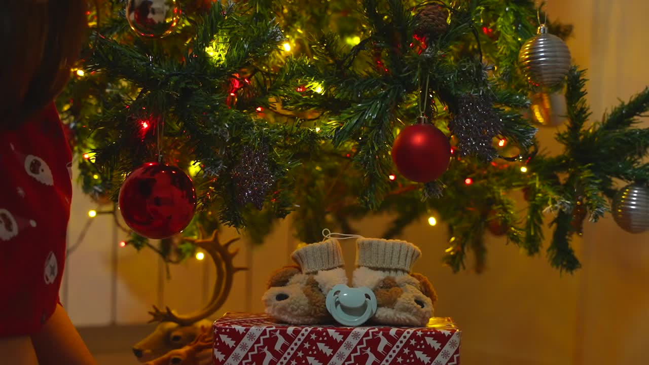 Close-up of baby booties on a wrapped present under the Christmas tree and female hands adding a pacifier as a baby announcement