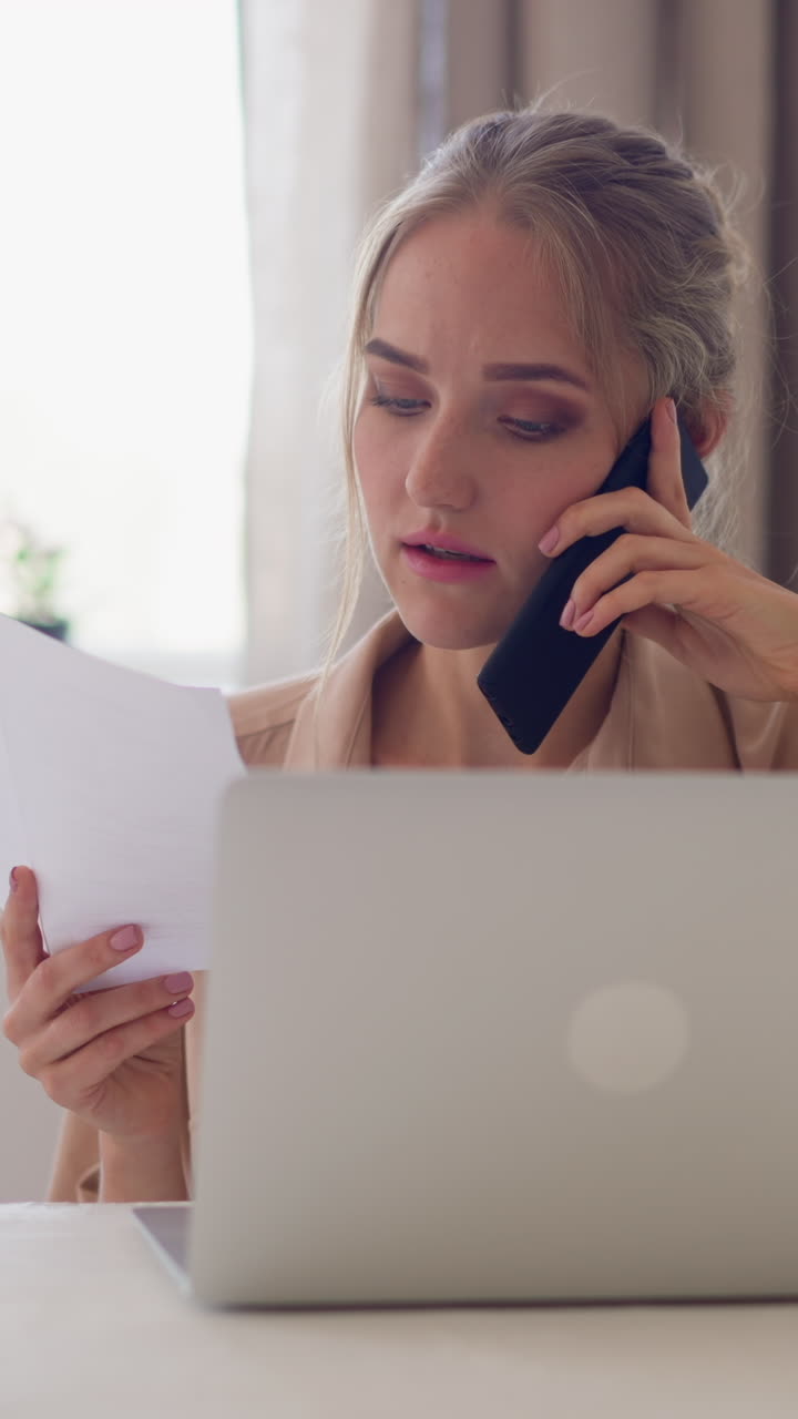Woman with blonde hair verifies data on paper via mobile phone sitting at table with laptop and coffee cup against bright window slow motion
