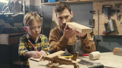 cheerful caucasian young father teaching small boy to work with timbers in workshop