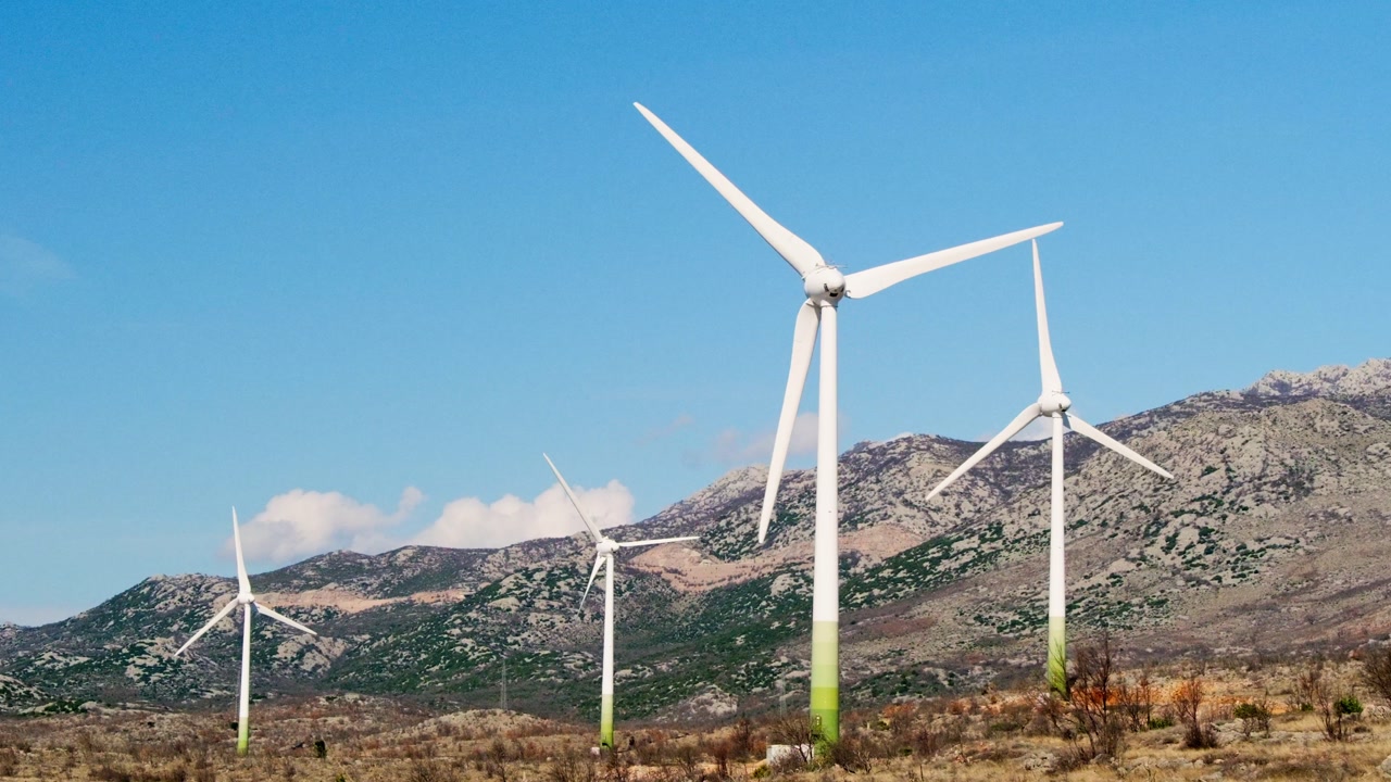 Wind turbines rotate at the foot of the Velebit mountains in Zadar County, producing renewable energy on a sunny day in a rugged, open landscape.