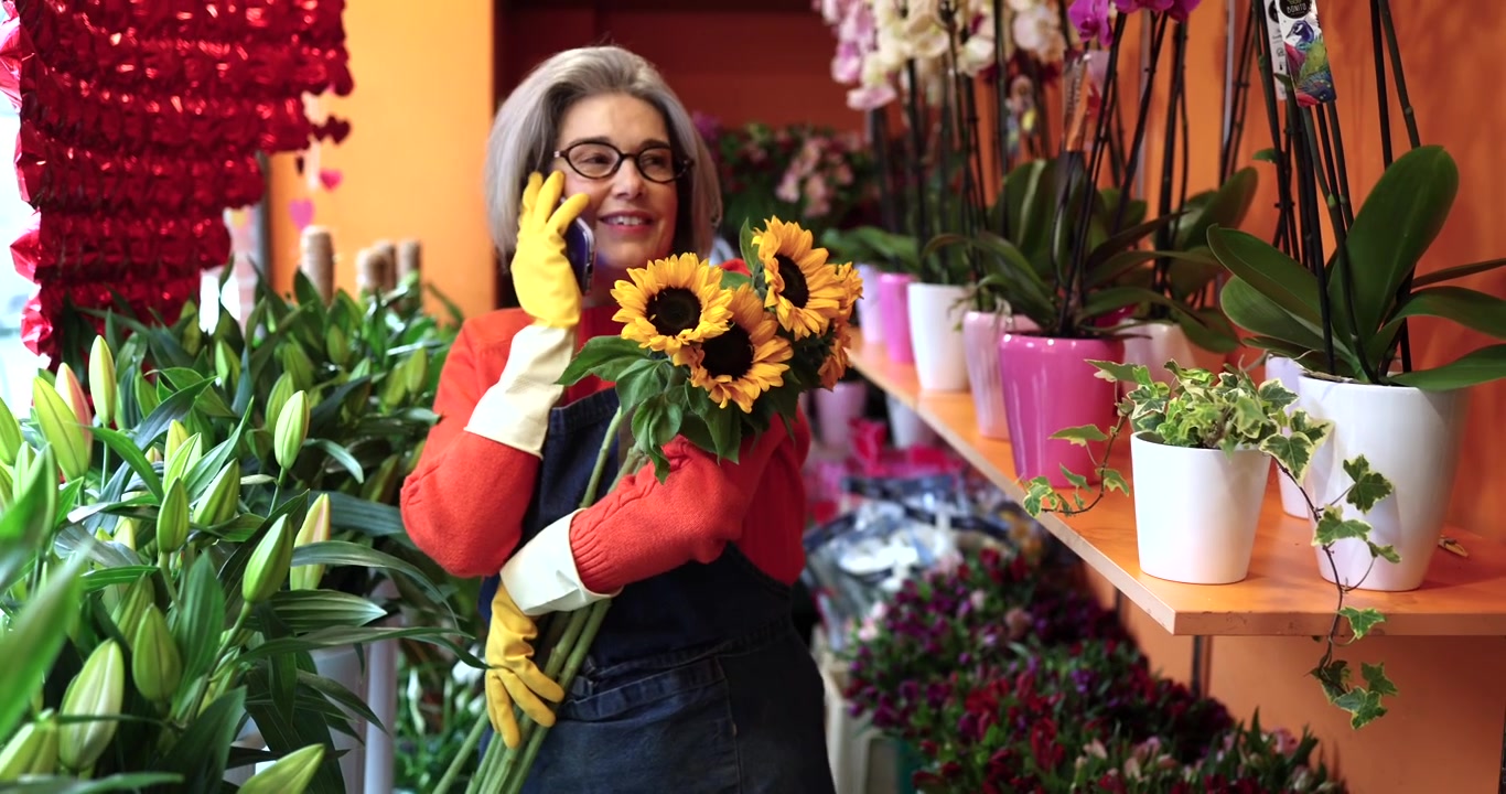 Fiorista anziano che parla al telefono mentre tiene dei girasoli in un negozio di fiori