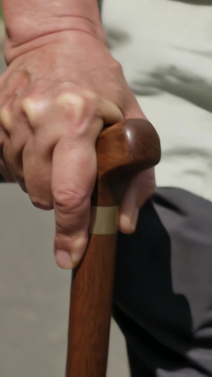 Video verticale: Anziano che sposta il peso, con la mano destra che impugna un bastone su un sentiero pedonale, indossando una camicia chiara.