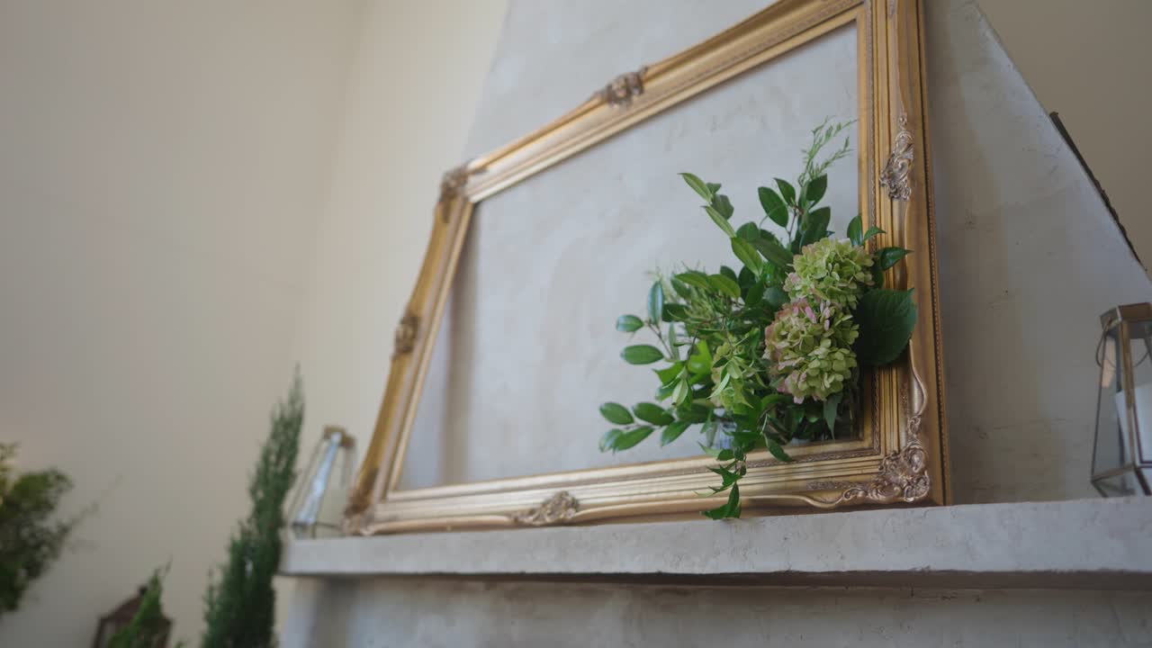 Una cornice dorata vuota, decorata con foglie verdi e fiori, è appoggiata alla parete sopra un camino classico, creando un tocco interno elegante, delicato e romantico.