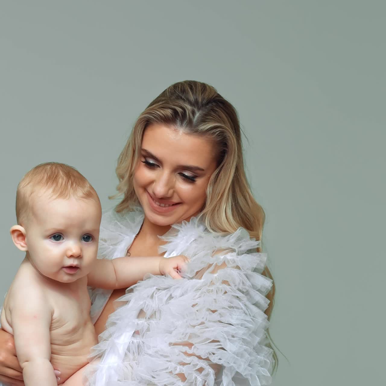 Wunderschöne, blonde, langhaarige Frau mit einem Baby im Arm. Die Mutter winkt und küsst das Kind. Weißer Hintergrund. Vertikaler Bildschirm