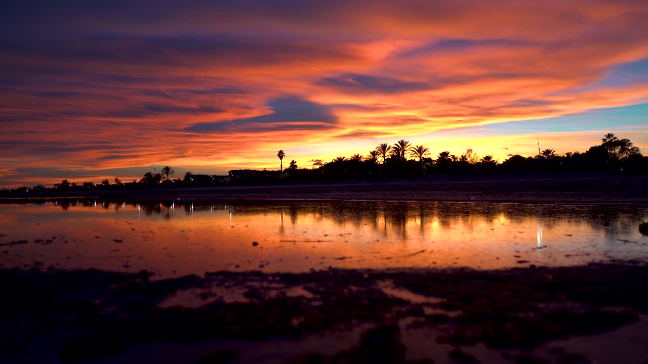 Time Lapse where burning clouds travel smoothly over the sky in an incredible sunrise, fairytale colors reflecting on the surface of the water in a beach lake, colorful movie scene